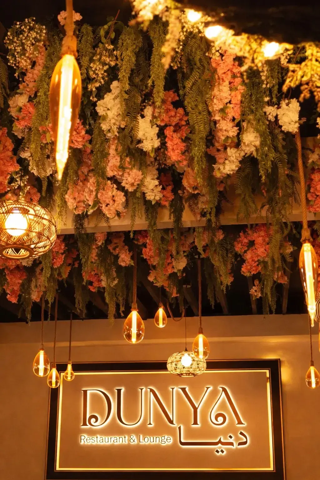 DUNYA illuminated sign beneath cascading florals and warm Edison bulbs